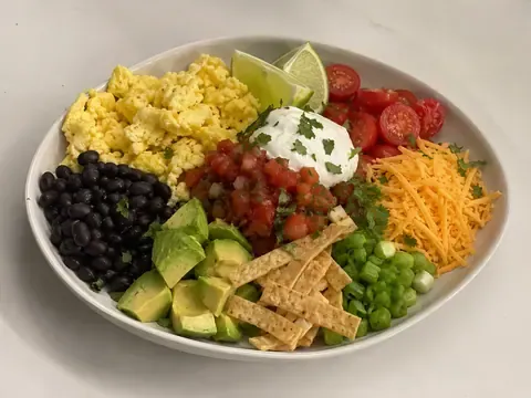Avocado egg breakfast taco bowl with scrambled eggs, black beans, avocado, salsa, tomatoes, cheese, yogurt, green onions, cilantro, tortilla strips, and lime.
