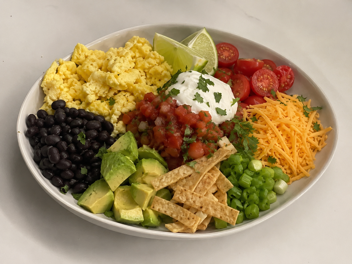 Avocado egg breakfast taco bowl with scrambled eggs, black beans, avocado, salsa, tomatoes, cheese, yogurt, green onions, cilantro, tortilla strips, and lime.