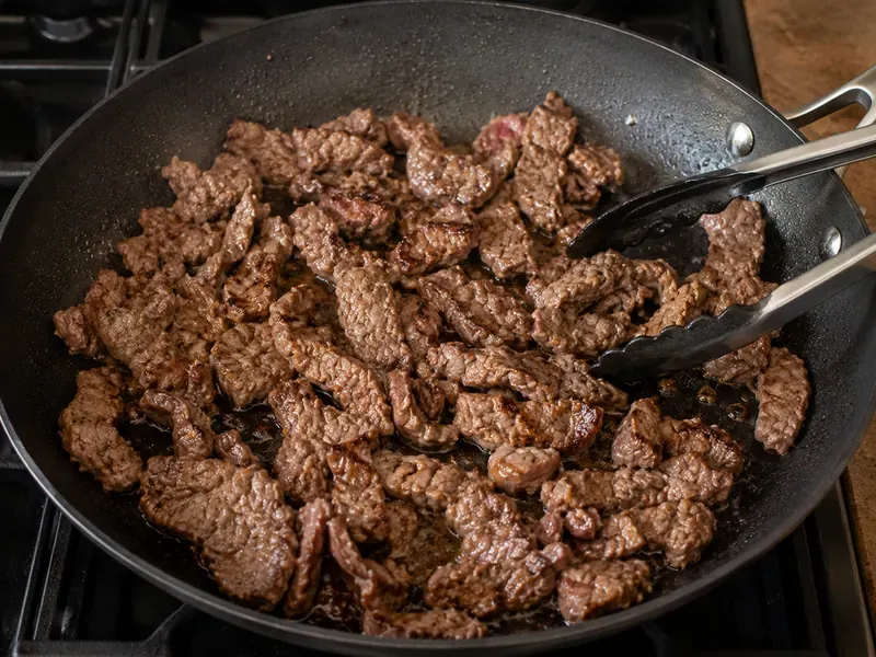 Thin slices of beef searing in a skillet until browned around the edges.