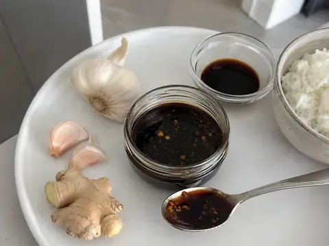 Blue Apron Soy Glaze Recipe final image showing a glossy dark soy glaze in a small jar or spoon, with garlic, ginger, soy sauce, rice bowl ingredients, and a realistic kitchen sauce setup.