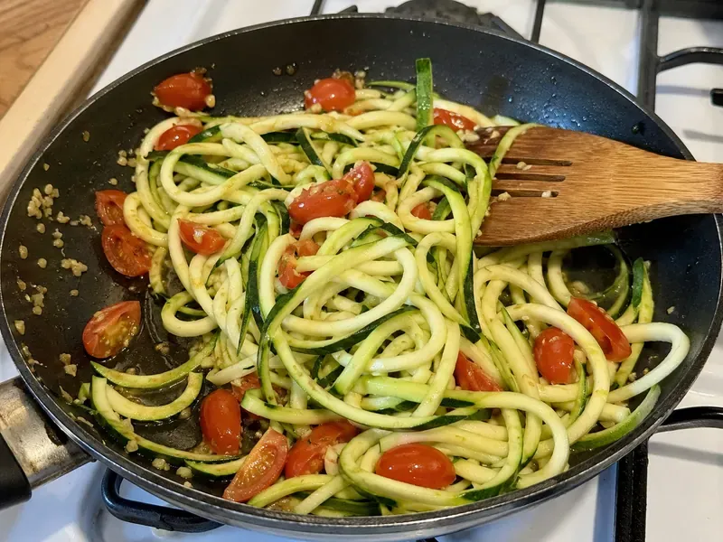 Zucchini noodles cooking with garlic and cherry tomatoes in a skillet.