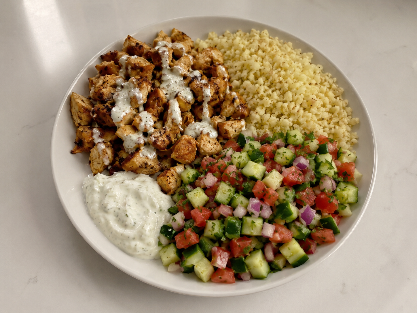 Chicken shawarma protein bowl with spiced chicken, cauliflower rice, cucumber tomato salad, parsley, and garlic yogurt sauce.