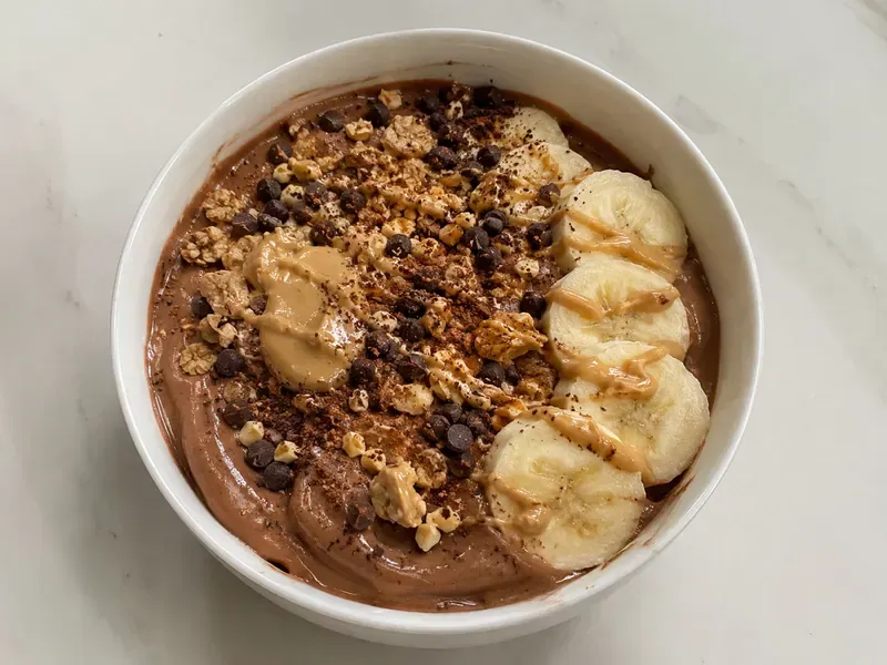 Chocolate peanut butter protein yogurt bowl with banana slices, chocolate chips, chopped peanuts, granola, and peanut butter drizzle.