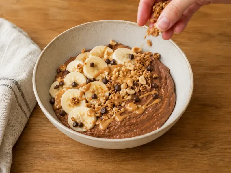 Chocolate protein yogurt bowl topped with banana, chocolate chips, chopped peanuts, granola, and peanut butter drizzle.