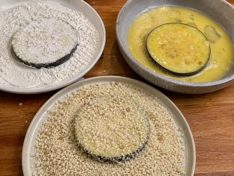 Eggplant rounds being coated in flour, egg, and breadcrumbs.
