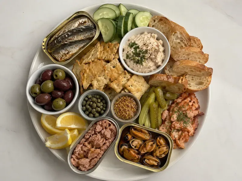 Easy tinned fish charcuterie board with sardines, tuna, crackers, pickles, olives, lemon, herbs, and creamy spread.