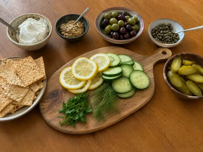 Lemon, cucumber, herbs, crackers, pickles, olives, capers, mustard, and creamy spread prepared for a snack board.
