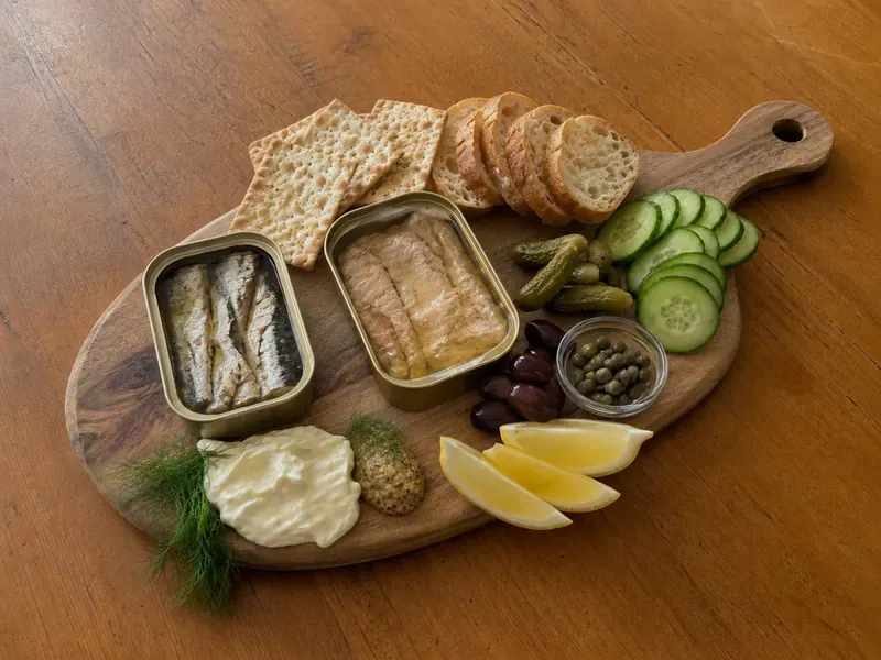 Tinned fish charcuterie board being arranged with opened tins, crackers, pickles, olives, lemon, and spreads.