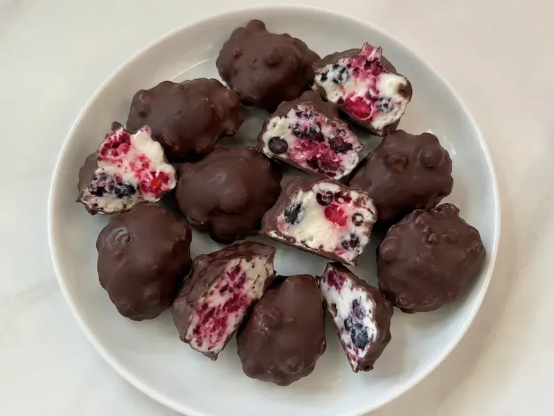 Frozen Yogurt Fruit Clusters final image showing bite-size frozen yogurt fruit clusters with berries inside and a crisp chocolate shell, some pieces cut open to show creamy yogurt and fruit, realistic freezer snack setup.