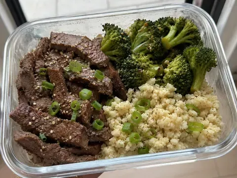 Garlic beef and broccoli meal prep box with beef slices, broccoli, cauliflower rice, green onions, and sesame seeds.