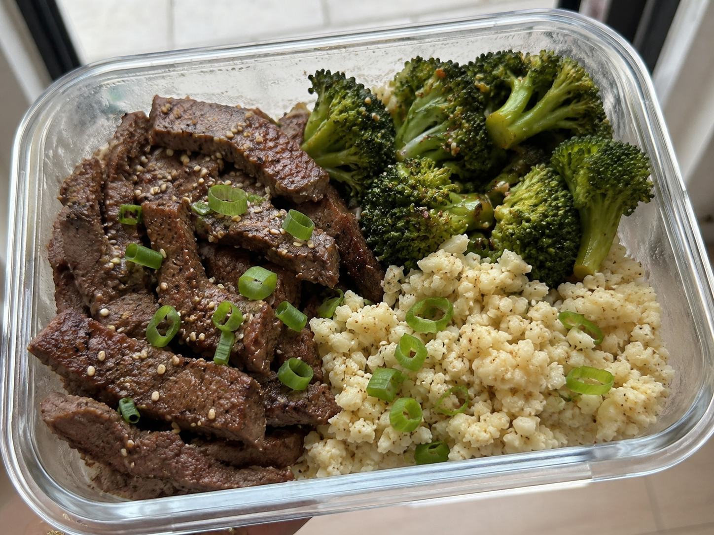 Garlic beef and broccoli meal prep box with beef slices, broccoli, cauliflower rice, green onions, and sesame seeds.