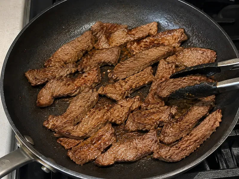 Thin slices of lean beef searing in a skillet until browned.