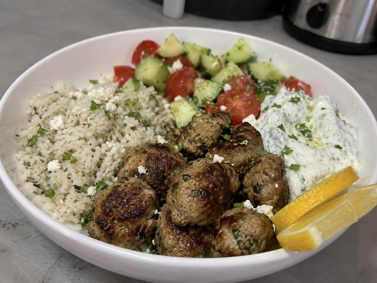 Greek turkey kofta bowl with herbed rice, cucumber tomato salad, tzatziki, feta, and lemon wedges.