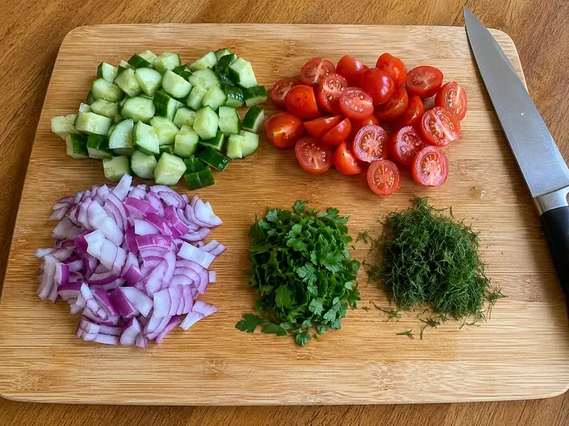 Diced cucumber, cherry tomatoes, red onion, parsley, and dill prepared for salad.
