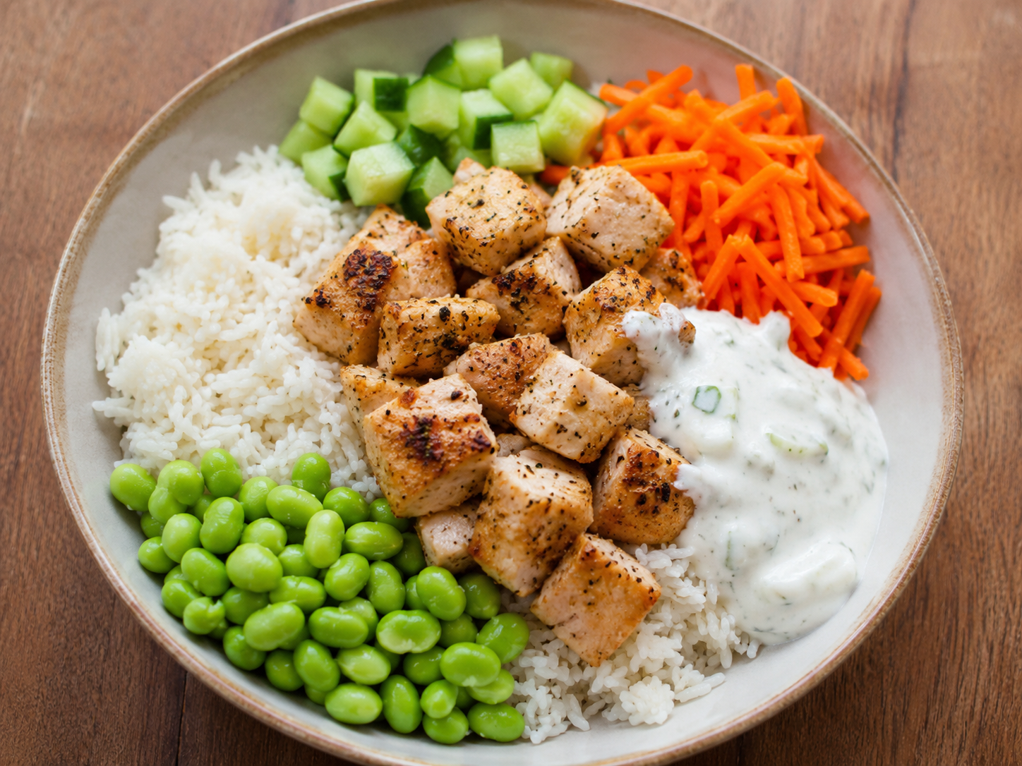 High protein chicken rice bowl with chicken breast, white rice, edamame, cucumber, carrots, and creamy sauce.