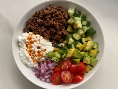 Hot honey cottage cheese beef bowl with seasoned ground beef, cottage cheese, cucumber, avocado, tomatoes, red onion, green onions, and hot honey drizzle.