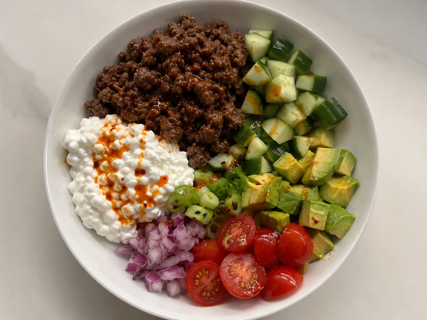 Hot honey cottage cheese beef bowl with seasoned ground beef, cottage cheese, cucumber, avocado, tomatoes, red onion, green onions, and hot honey drizzle.