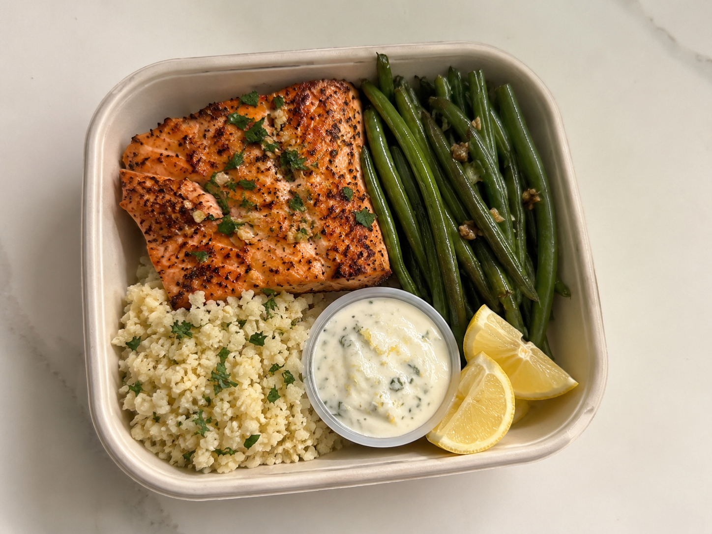 Lemon garlic salmon meal prep box with green beans, cauliflower rice, lemon wedges, parsley, and yogurt sauce.