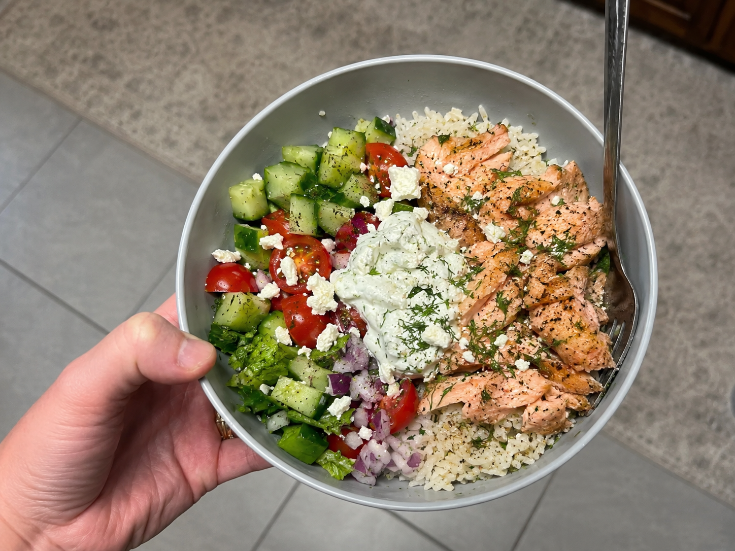 Lemon herb salmon bowl with rice, greens, cucumber, tomatoes, red onion, feta, herbs, and yogurt sauce.