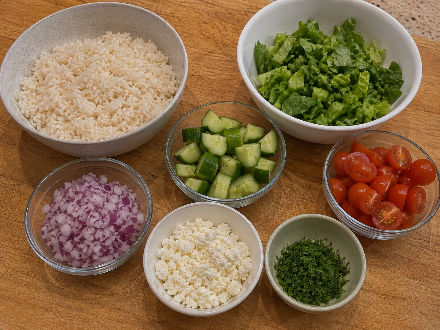 Rice, greens, cucumber, tomatoes, red onion, feta, and herbs prepared for a salmon bowl.