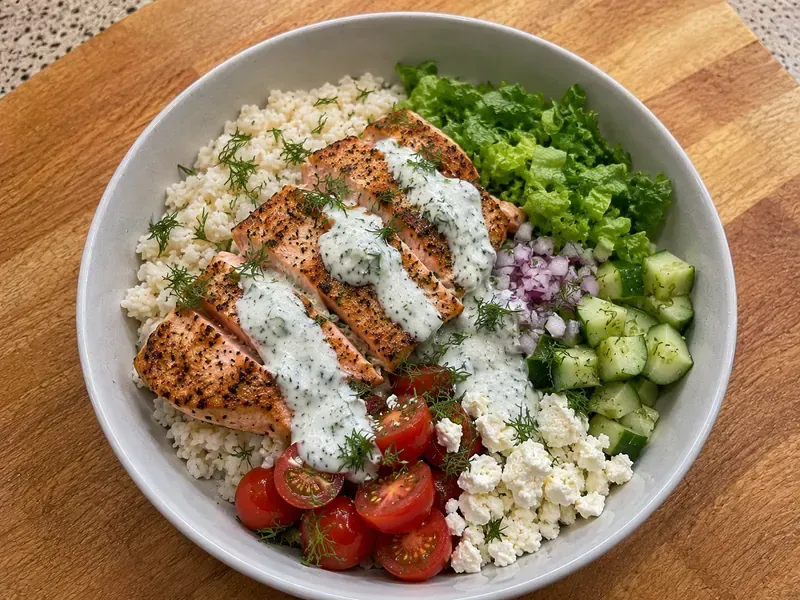 Finished lemon herb salmon bowl with salmon, rice, greens, cucumber, tomatoes, red onion, feta, herbs, and yogurt sauce.