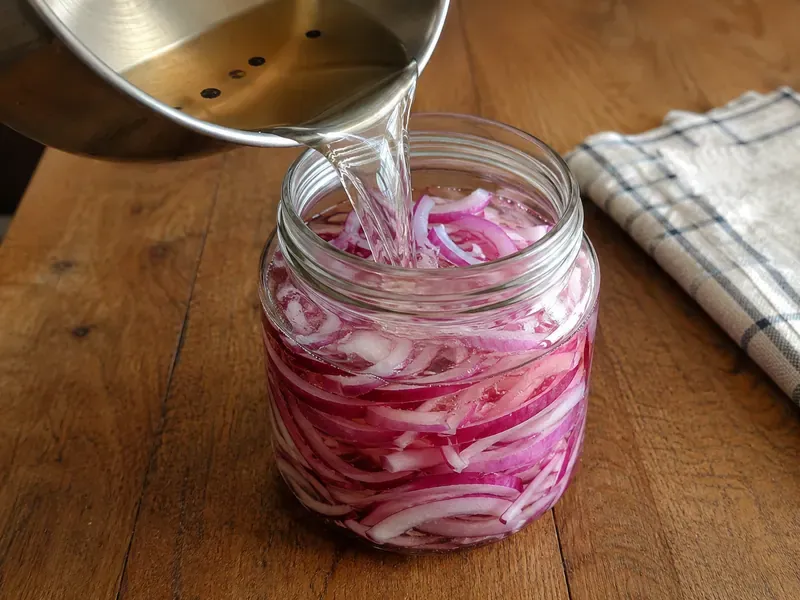 Warm vinegar brine poured over sliced red onions in a glass jar.