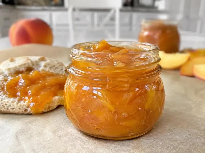 Small-Batch Peach Jam in a glass jar with a spoon and fresh peaches nearby.