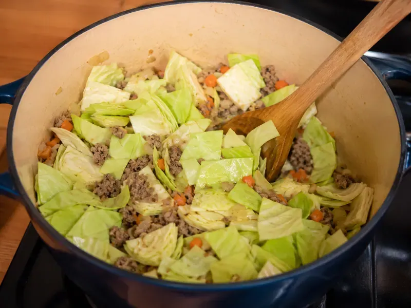 Chopped cabbage stirred into a pot with ground beef, onion, garlic, and carrot.