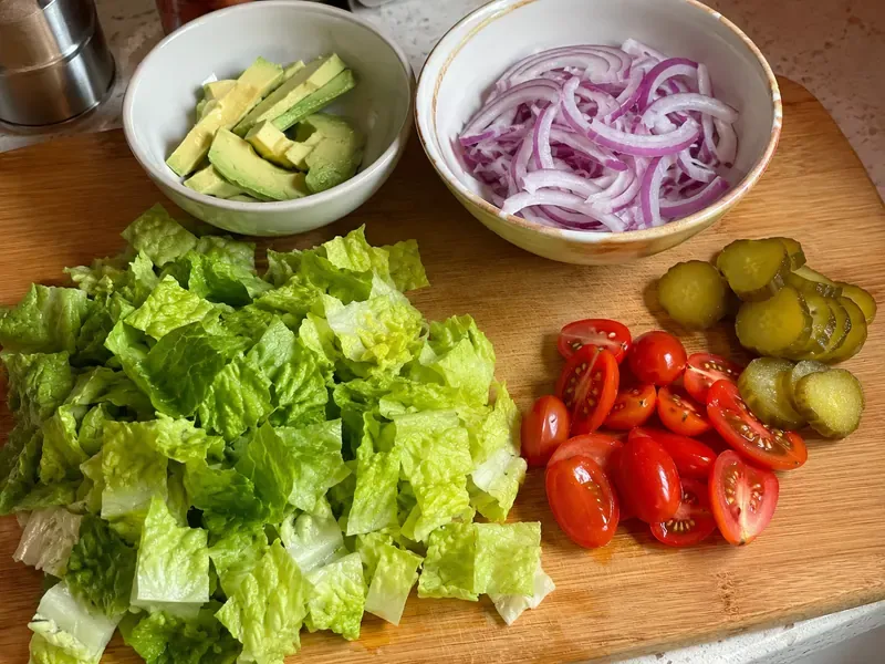 Lettuce, tomatoes, pickles, red onion, and avocado prepared for a turkey burger protein bowl.
