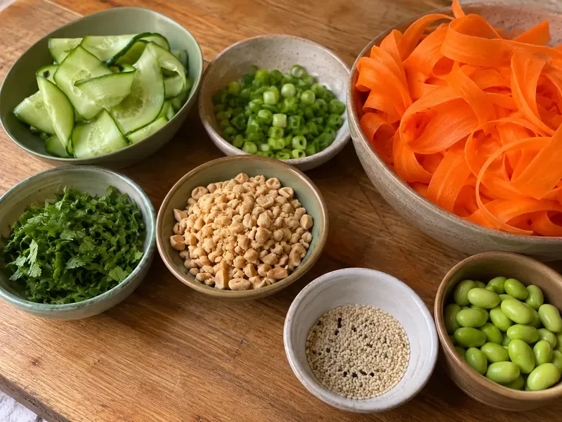 Cucumber, green onions, cilantro, peanuts, sesame seeds, edamame, and carrot ribbons prepared for salad.